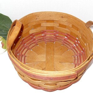 May include: A round, woven basket with two leather handles. The basket is made of natural wood with a red trim around the top. The basket is empty.