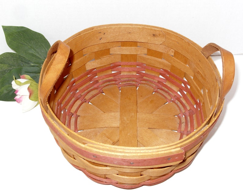 May include: A round, woven basket with two leather handles. The basket is made of natural wood with a red trim around the top. The basket is empty.