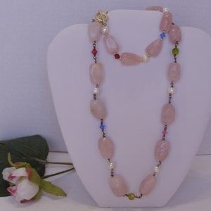 May include: A long necklace with large, light pink, oval-shaped beads. The necklace is accented with small white pearls and colorful crystals. It has a gold-colored clasp and is displayed on a white mannequin.
