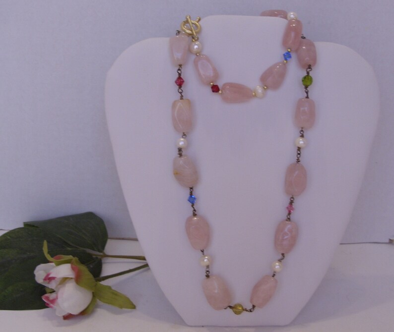 May include: A long necklace with large, light pink, oval-shaped beads. The necklace is accented with small white pearls and colorful crystals. It has a gold-colored clasp and is displayed on a white mannequin.