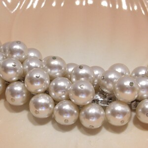May include: A vintage pearl bracelet with multiple strands of large, round, off-white pearls. The pearls are connected by a silver-toned metal chain and clasp. The bracelet is displayed on a light peach-colored surface.