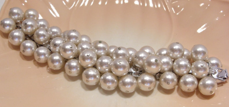 May include: A vintage pearl bracelet with multiple strands of large, round, off-white pearls. The pearls are connected by a silver-toned metal chain and clasp. The bracelet is displayed on a light peach-colored surface.