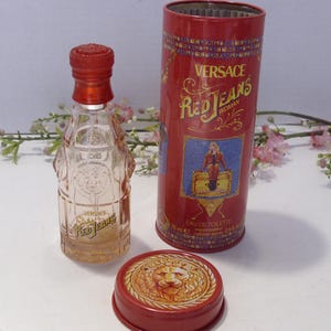 May include: A clear glass perfume bottle with a red cap and the text Versace Red Jeans, accompanied by a matching red cylindrical metal tin featuring a graphic illustration and the text Versace Red Jeans Woman, plus a separate red metal lid with a lion.