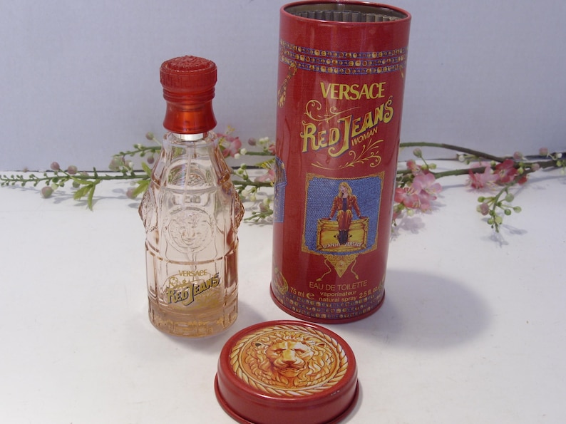 May include: A clear glass perfume bottle with a red cap and the text Versace Red Jeans, accompanied by a matching red cylindrical metal tin featuring a graphic illustration and the text Versace Red Jeans Woman, plus a separate red metal lid with a lion.