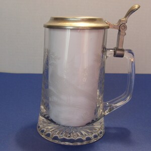 May include: Clear glass beer stein with a hinged metal lid and handle. The glass features an etched design of a bird and foliage. The base of the stein has a decorative, textured pattern.