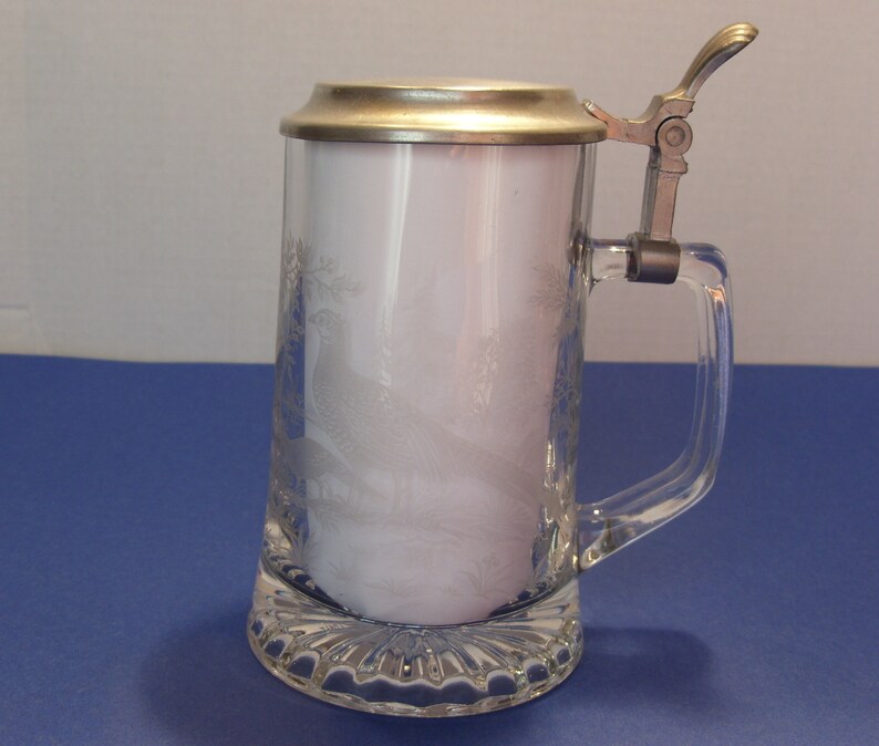 May include: Clear glass beer stein with a hinged metal lid and handle. The glass features an etched design of a bird and foliage. The base of the stein has a decorative, textured pattern.