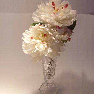 Puede incluir: Un jarr&oacute;n de cristal transparente contiene un ramo de peon&iacute;as blancas con detalles rojos y amarillos. El jarr&oacute;n tiene una base hexagonal y un cuerpo texturizado con forma de diamante. Las flores est&aacute;n en plena floraci&oacute;n, creando un arreglo floral delicado.