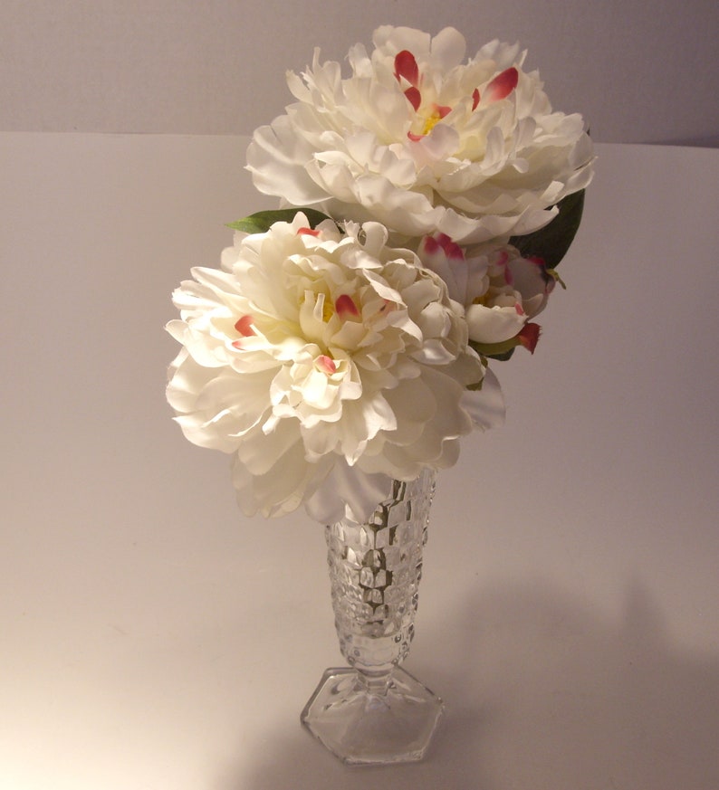Puede incluir: Un jarr&oacute;n de cristal transparente contiene un ramo de peon&iacute;as blancas con detalles rojos y amarillos. El jarr&oacute;n tiene una base hexagonal y un cuerpo texturizado con forma de diamante. Las flores est&aacute;n en plena floraci&oacute;n, creando un arreglo floral delicado.