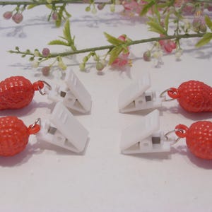 Op de afbeelding: Zes witte wasknijpers met rode, getextureerde aardbei-vormige bedels. De clips liggen op een wit oppervlak, met een takje roze en groene bloemen op de achtergrond.