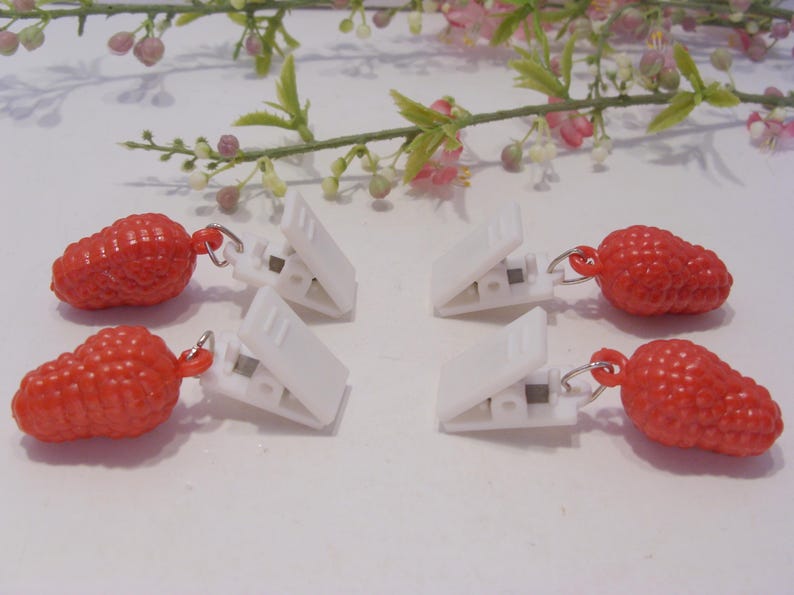 Op de afbeelding: Zes witte wasknijpers met rode, getextureerde aardbei-vormige bedels. De clips liggen op een wit oppervlak, met een takje roze en groene bloemen op de achtergrond.