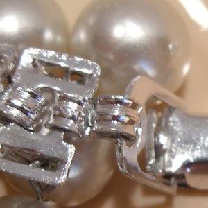 May include: Close-up of a silver-toned clasp, likely from a necklace or bracelet, with a series of interlocking rectangular pieces. The clasp is set against a backdrop of large, round, off-white pearls. The word "JAPAN" is visible on the clasp.
