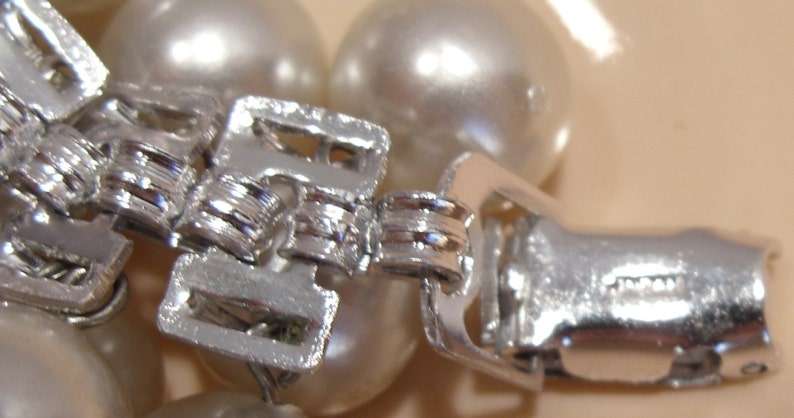 May include: Close-up of a silver-toned clasp, likely from a necklace or bracelet, with a series of interlocking rectangular pieces. The clasp is set against a backdrop of large, round, off-white pearls. The word "JAPAN" is visible on the clasp.