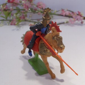 May include: A plastic toy knight on horseback, the knight is wearing black armor with a blue and gold helmet, the horse is gold with red and gold accents, the knight is holding a red and orange spear.