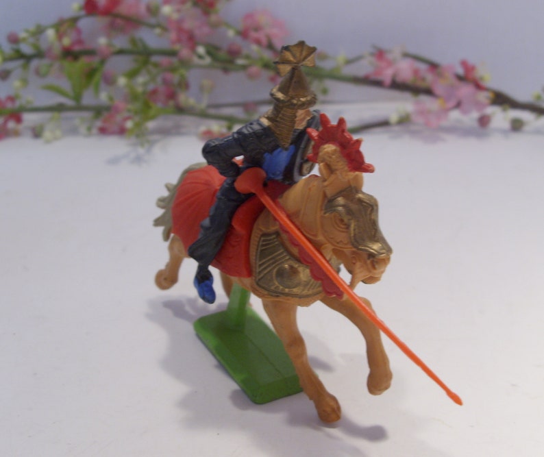 May include: A plastic toy knight on horseback, the knight is wearing black armor with a blue and gold helmet, the horse is gold with red and gold accents, the knight is holding a red and orange spear.