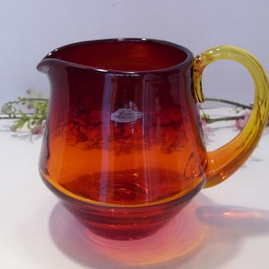 May include: A glass pitcher with a vibrant gradient of red to orange hues. The handle is a bright yellow, contrasting with the body. The pitcher has a rounded shape and a pouring spout. A small label is visible on the front.