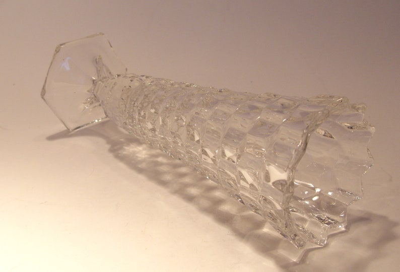 Puede incluir: Jarr&oacute;n de vidrio transparente con un cuerpo texturizado con dise&ntilde;o de diamantes y una base hexagonal. El jarr&oacute;n cil&iacute;ndrico est&aacute; inclinado, mostrando su intrincado dise&ntilde;o. El vidrio refleja la luz, resaltando su transparencia y atractivo decorativo.