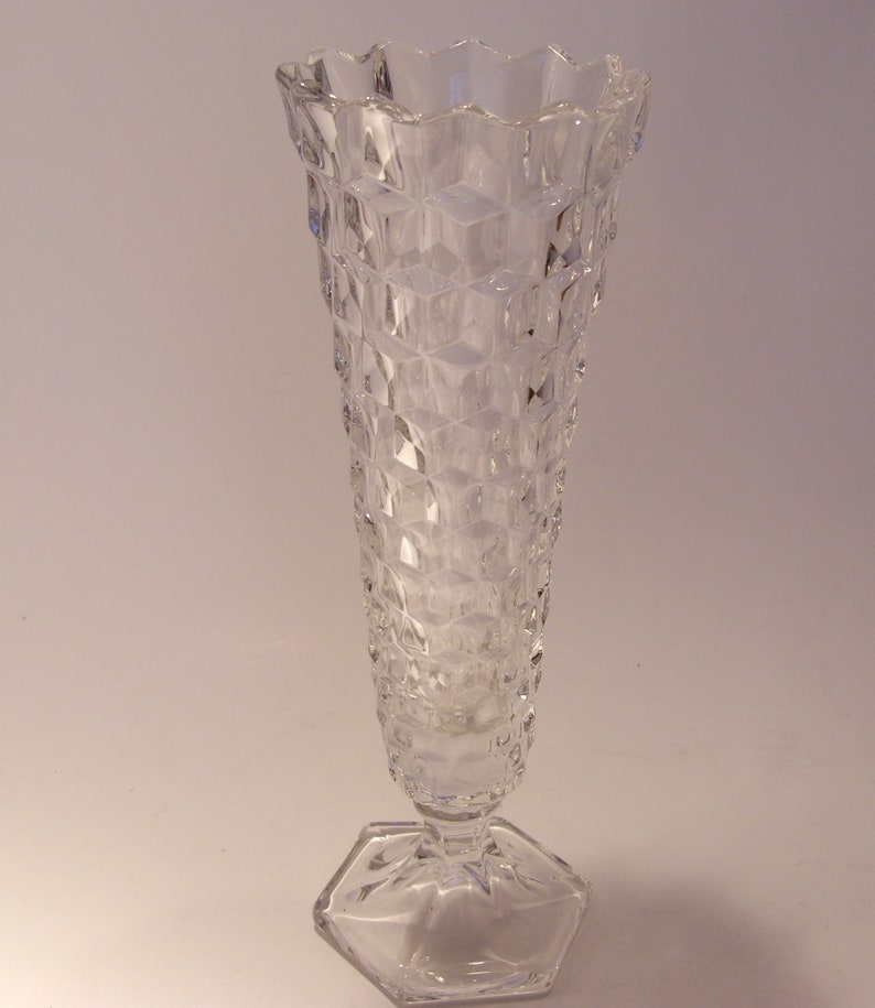 Puede incluir: Un jarr&oacute;n de cristal transparente con un patr&oacute;n de diamantes y una base hexagonal. El jarr&oacute;n tiene un borde acampanado y festoneado y un cuerpo largo y esbelto. El jarr&oacute;n mide unos 25 cm de alto.