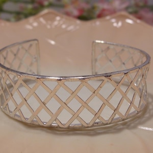 May include: Silver-toned cuff bracelet with an open, lattice-like design. The bracelet features a series of intersecting lines forming a diamond pattern. The open design allows light to pass through, creating a delicate and airy aesthetic. The bracelet is displayed on a light-colored surface.