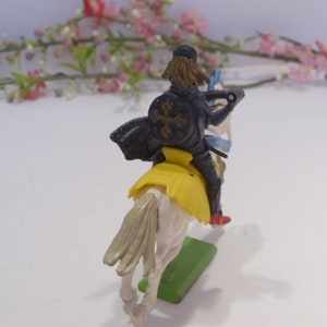 May include: A plastic toy knight riding a white horse. The knight is wearing black armor with a yellow sash and a black cross on his shield. The horse has a yellow saddle and a blue and white tail.