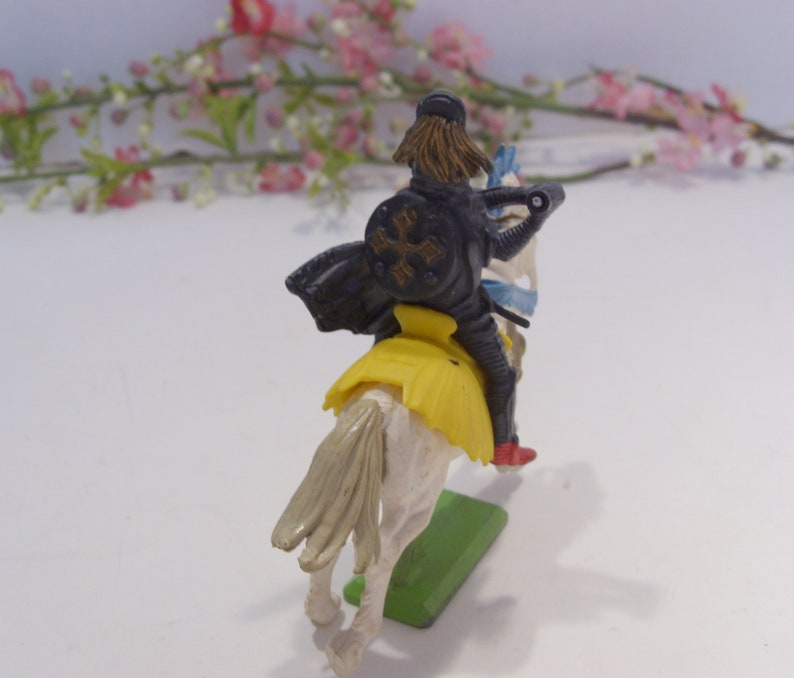 May include: A plastic toy knight riding a white horse. The knight is wearing black armor with a yellow sash and a black cross on his shield. The horse has a yellow saddle and a blue and white tail.