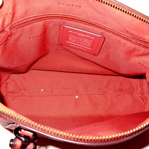 May include: Open view of a red handbag interior with a zippered pocket and a small leather tag. The interior is lined with red fabric and features a gold zipper and hardware.