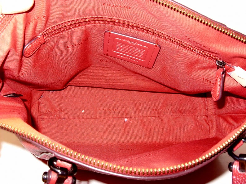 May include: Open view of a red handbag interior with a zippered pocket and a small leather tag. The interior is lined with red fabric and features a gold zipper and hardware.