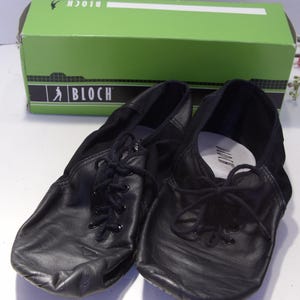Puede incluir: Un par de zapatillas de ballet de cuero negro con cordones, frente a una caja verde con el logo de Bloch. Los zapatos tienen un dise&ntilde;o suave y flexible, ideal para bailar.