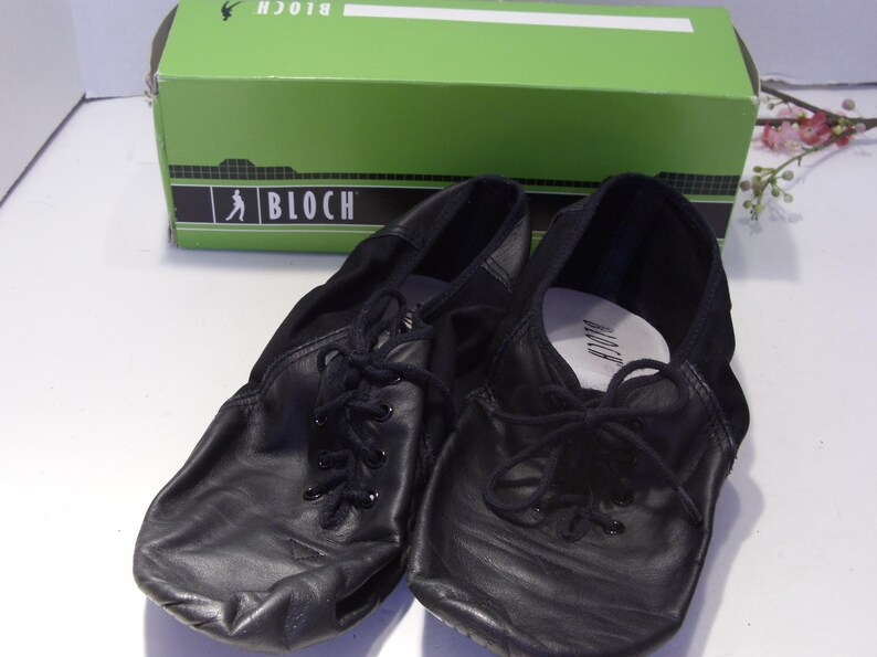 Puede incluir: Un par de zapatillas de ballet de cuero negro con cordones, frente a una caja verde con el logo de Bloch. Los zapatos tienen un dise&ntilde;o suave y flexible, ideal para bailar.