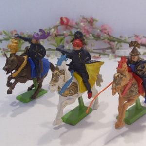 May include: Three plastic toy knights on horseback, each wearing a different colored helmet and armor. The knights are riding white, brown, and black horses. The horses are all in a galloping pose.