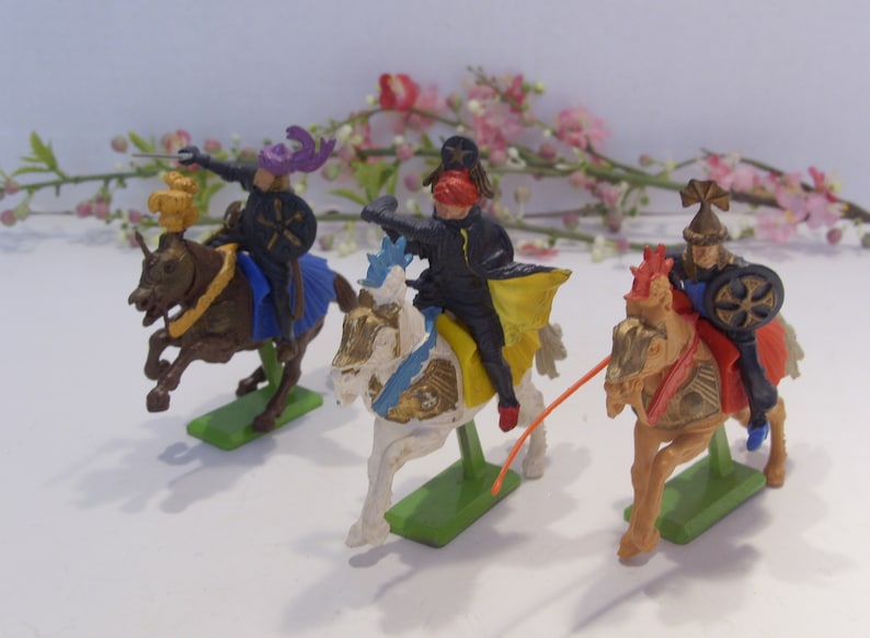 May include: Three plastic toy knights on horseback, each wearing a different colored helmet and armor. The knights are riding white, brown, and black horses. The horses are all in a galloping pose.