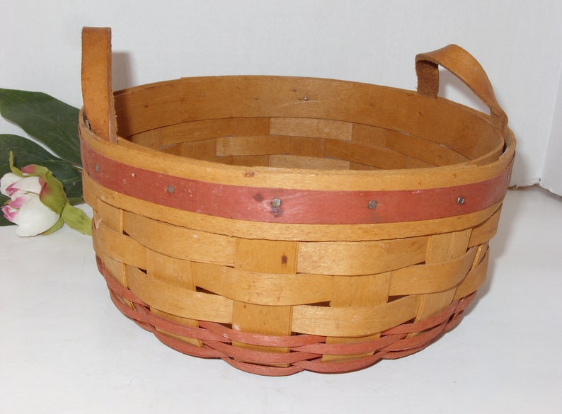 May include: A woven basket with two leather handles. The basket is made of natural wood and has a red trim around the top and bottom.