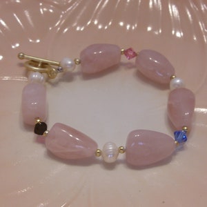 May include: A bracelet featuring large, polished rose quartz beads, interspersed with small pearls and gold-colored accents. The bracelet has a toggle clasp and includes small pink and blue crystal beads.