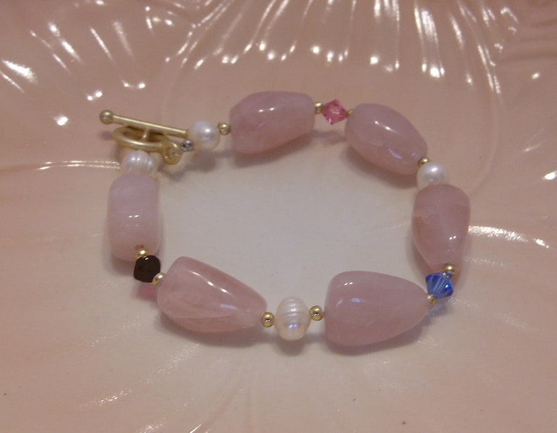 May include: A bracelet featuring large, polished rose quartz beads, interspersed with small pearls and gold-colored accents. The bracelet has a toggle clasp and includes small pink and blue crystal beads.