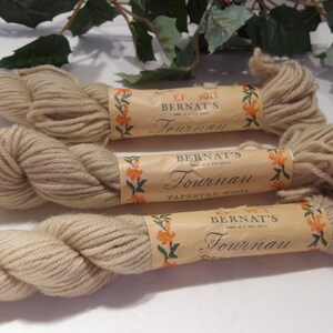 May include: Three skeins of beige Bernat's Tournau Tapestry Wool yarn. Each skein is wrapped in a paper label with floral accents and the brand name. The yarn is a light, neutral color, suitable for various crafting projects.