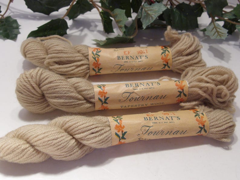 May include: Three skeins of beige Bernat's Tournau Tapestry Wool yarn. Each skein is wrapped in a paper label with floral accents and the brand name. The yarn is a light, neutral color, suitable for various crafting projects.