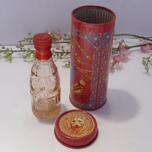 May include: A clear glass perfume bottle with a red cap stands next to a cylindrical red metal container. The container features gold and blue decorative patterns and the text VERSACE PROFUMI. A separate red lid displays a gold lion head emblem.