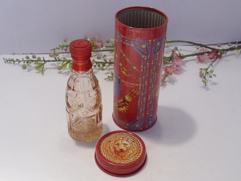 May include: A clear glass perfume bottle with a red cap stands next to a cylindrical red metal container. The container features gold and blue decorative patterns and the text VERSACE PROFUMI. A separate red lid displays a gold lion head emblem.