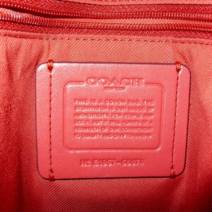 May include: Close-up of the interior of a red Coach bag. A rectangular red leather tag is stitched inside, bearing the Coach logo and text. The bag's lining and zipper are also red.