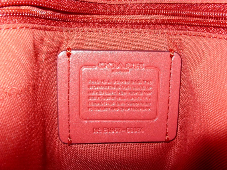 May include: Close-up of the interior of a red Coach bag. A rectangular red leather tag is stitched inside, bearing the Coach logo and text. The bag's lining and zipper are also red.