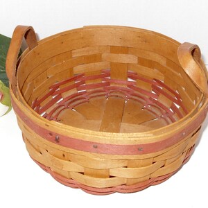 May include: A round, woven basket with two leather handles. The basket is made of natural wood with a red trim around the top and bottom.