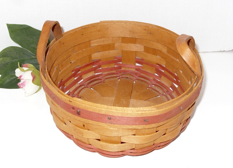 May include: A round, woven basket with two leather handles. The basket is made of natural wood with a red trim around the top and bottom.