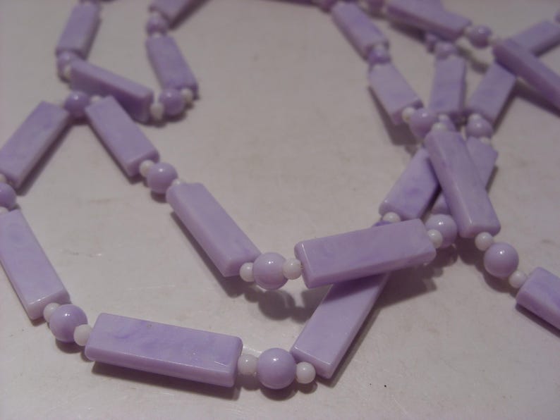 May include: A long necklace featuring a repeating pattern of light purple rectangular beads and smaller light purple spherical beads, separated by tiny white spacer beads. The rectangular beads have a marbled texture in shades of lavender.