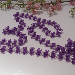 May include: A long beaded necklace featuring a repeating pattern of small, translucent purple flower-shaped beads alternating with clusters of tiny, round, opaque white beads. The necklace is arranged in a loose, winding shape on a white surface.