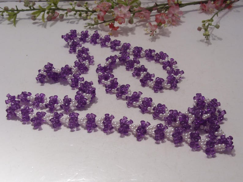 May include: A long beaded necklace featuring a repeating pattern of small, translucent purple flower-shaped beads alternating with clusters of tiny, round, opaque white beads. The necklace is arranged in a loose, winding shape on a white surface.