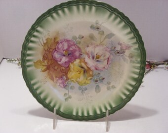 May include: A vintage porcelain plate with a green scalloped edge and a floral design featuring pink, yellow, and white flowers. The flowers are painted in a realistic style and are surrounded by green leaves.
