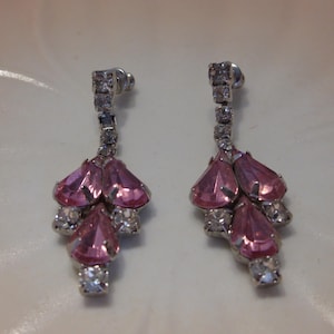 Peut inclure: Une paire de boucles d'oreilles pendantes avec une monture en métal argenté. Chaque boucle d'oreille présente une série de strass clairs taillés en carré menant à un groupe de pierres roses en forme de goutte. Les boucles d'oreilles sont percées.