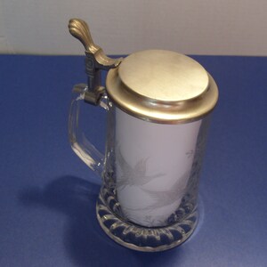 May include: A clear glass beer stein with a silver-colored metal lid and handle. The stein features an etched design of a bird. The base of the stein has a decorative, scalloped edge.