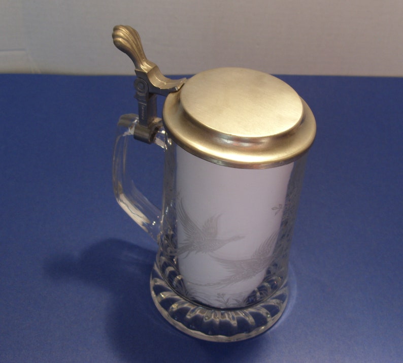 May include: A clear glass beer stein with a silver-colored metal lid and handle. The stein features an etched design of a bird. The base of the stein has a decorative, scalloped edge.