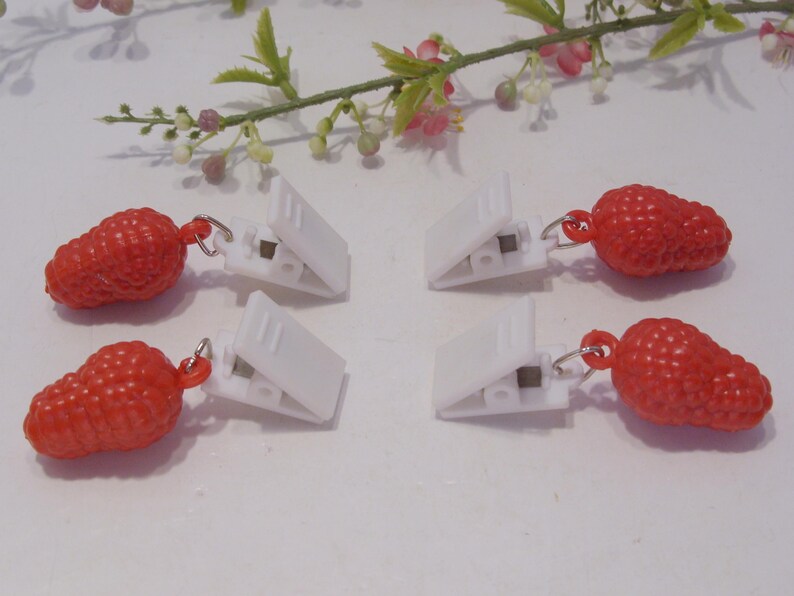 Op de afbeelding: Vier witte plastic clips met rode aardbei-vormige bedels. De bedels zijn met kleine zilverkleurige ringen aan de clips bevestigd. Een takje bloemen met roze en witte bloesems op de achtergrond.