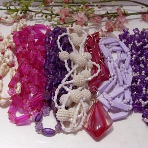 May include: A collection of six necklaces featuring various designs: a white seashell strand, a bright pink rectangular bead strand, a deep purple bead strand, a white textured bead strand, a light lavender rectangular bead strand, and a purple beaded strand with a large red diamond-shaped pendant.